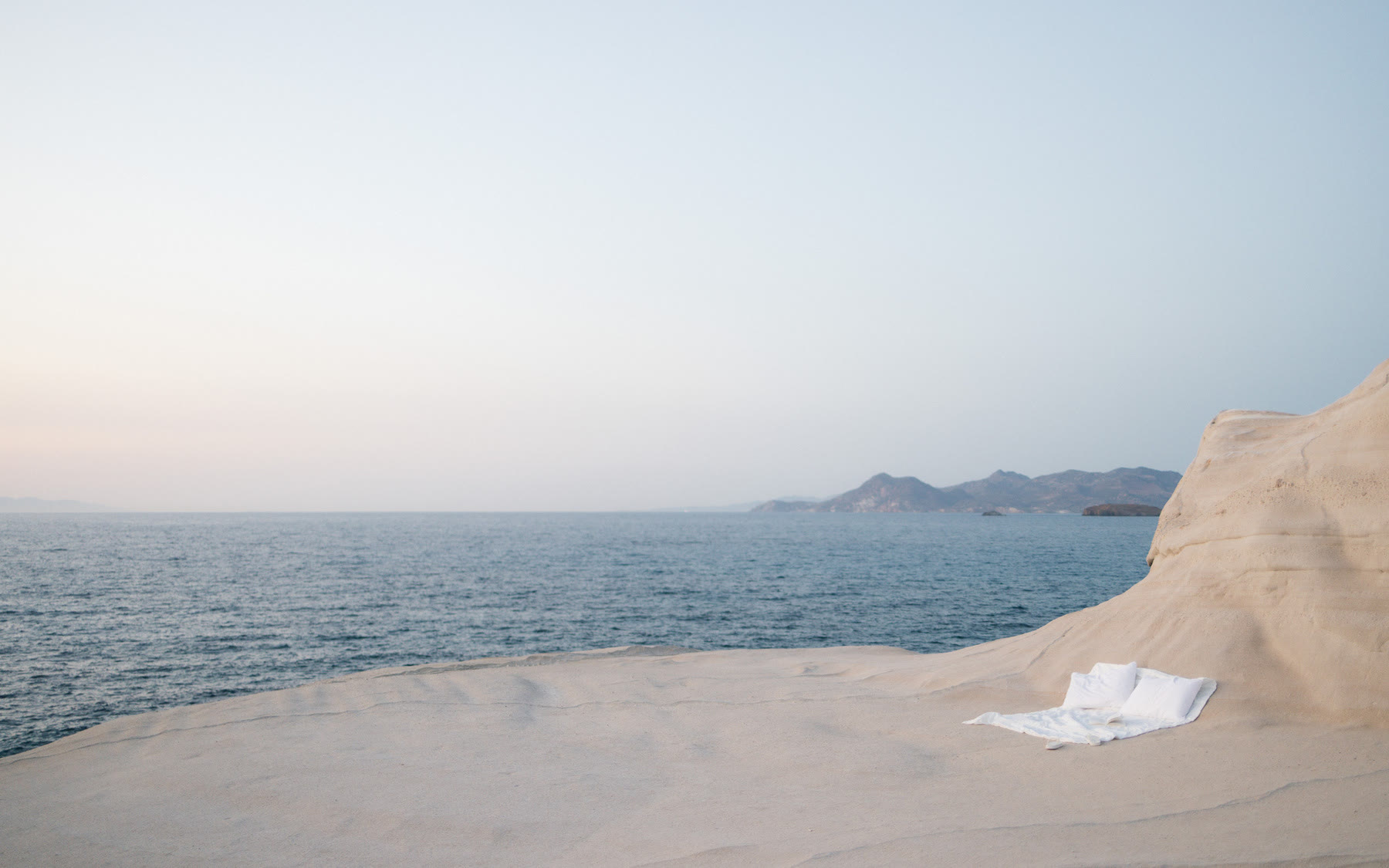 A serene coastal scene on the Greek Island of Milos features a minimalist setup with a sheet and pillow on a rocky shoreline overlooking calm waters and distant islands.